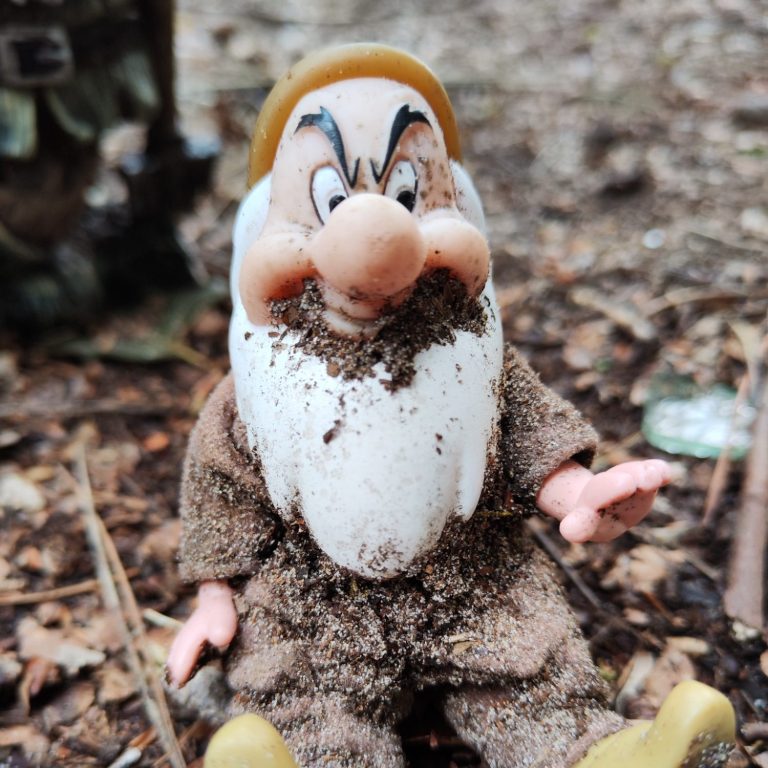 Een vieze, zandbedekte dwerg met een grote baard en een gele hoed, zittend op de grond.